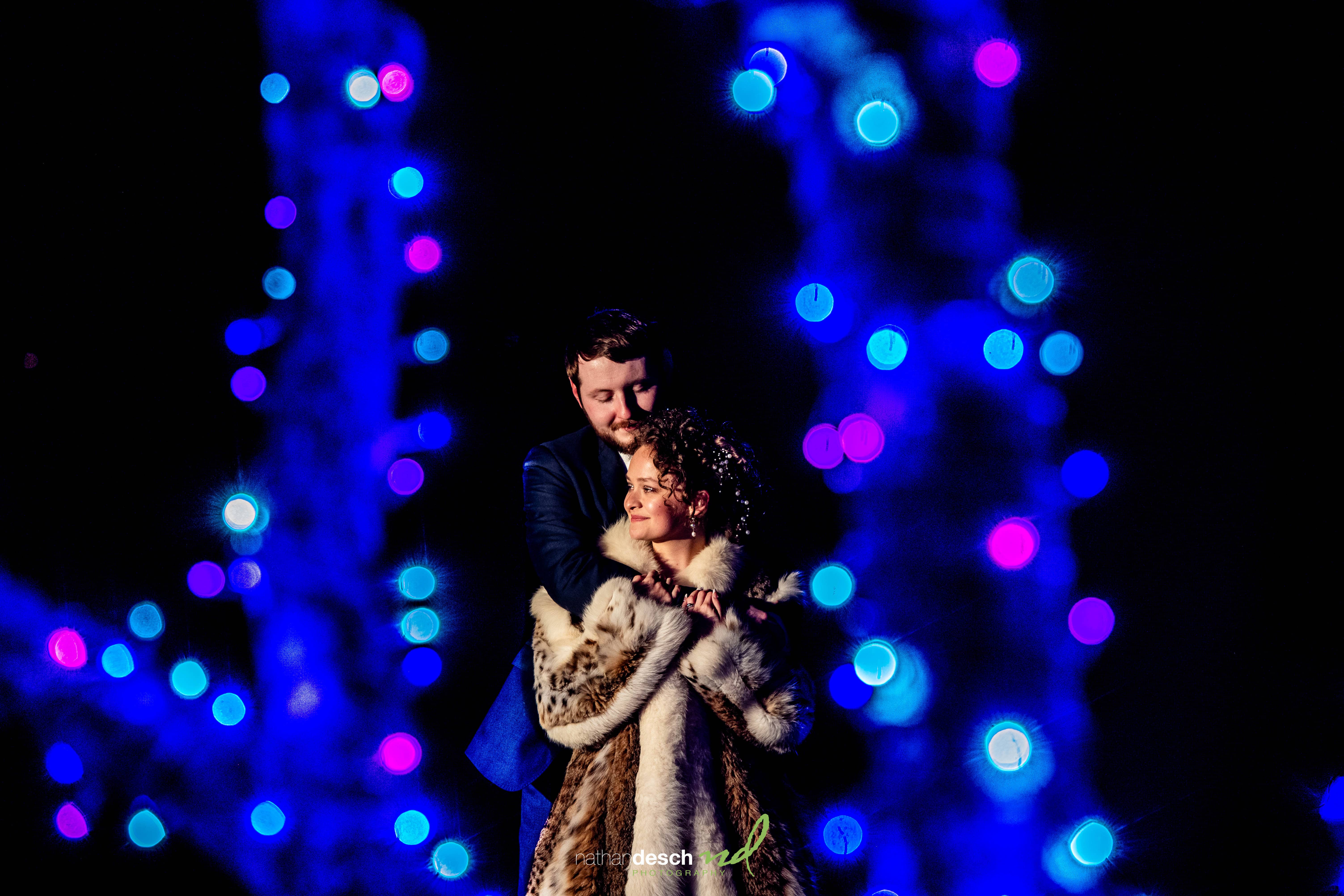 Bride and groom hugging in a fur coat with blue and purple bokeh lights, romantic wedding photography moment.